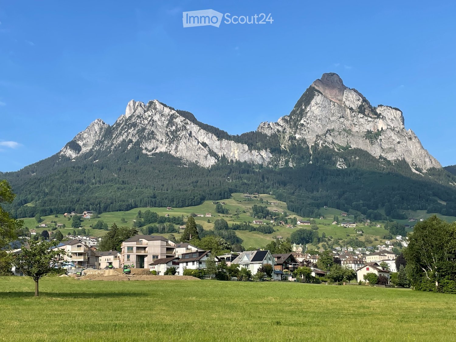 A picturesque village nestled in a lush green valley with rolling hills and a prominent mountain range in the background. The village features scattered houses and buildings surrounded by trees and open fields.