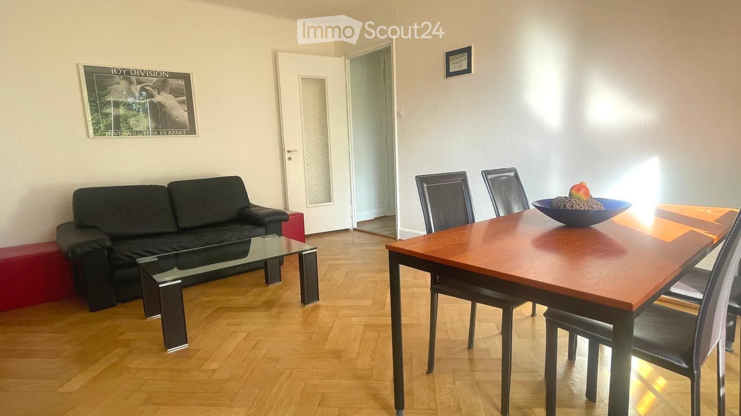 A modern living and dining area with a black sofa, glass coffee table, wooden dining table, and four chairs. A bowl with fruit sits on the dining table, and a framed poster adorns the wall.