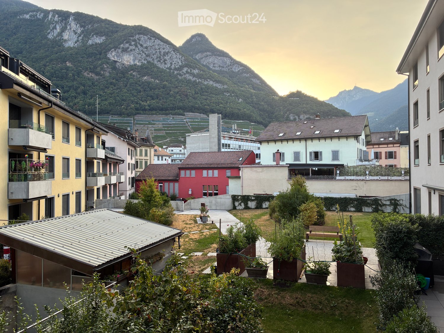 A serene garden area with potted plants, a pathway, and a view of a town with colorful buildings and distant mountains.