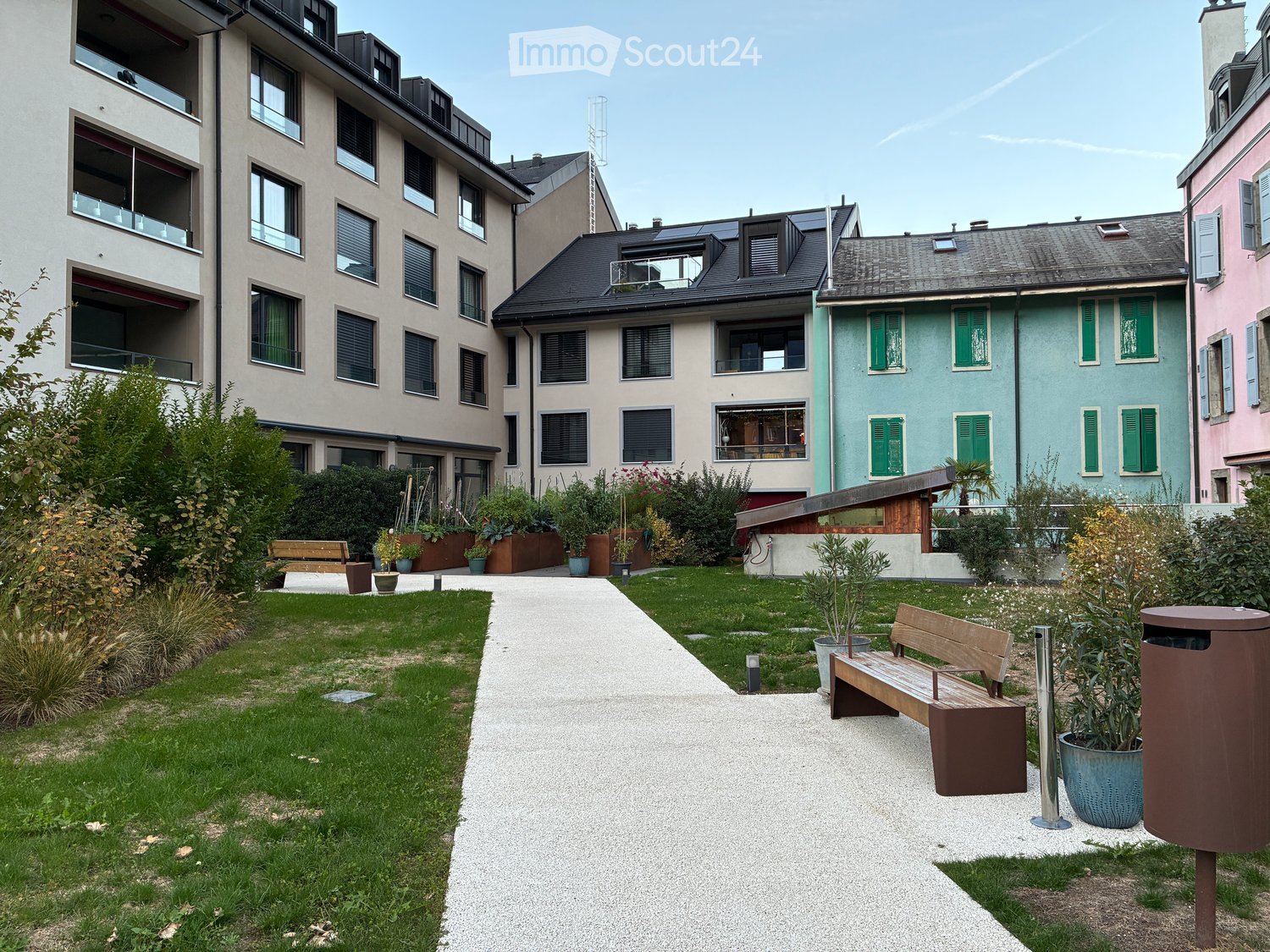 Courtyard with a paved pathway, grassy areas, benches, potted plants, and surrounding residential buildings.