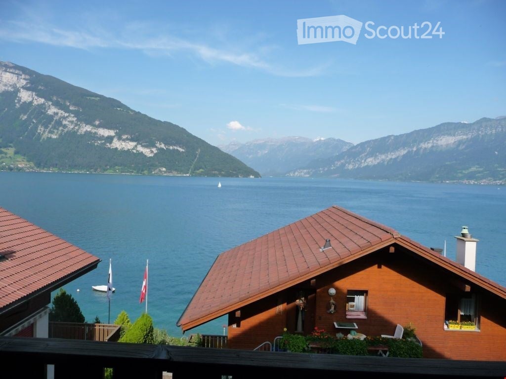 A cozy wooden house with a red-tiled roof, situated on a balcony overlooking a serene lake and distant mountains.