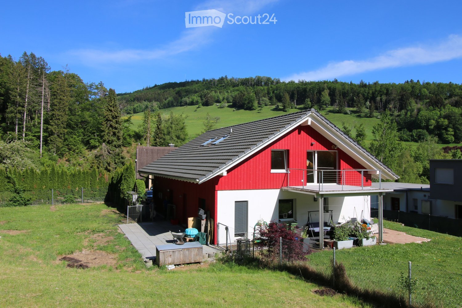 A two-story house with red and white exterior, grey roof, and a small balcony. The house is surrounded by a well-maintained garden with various plants and trees, and is situated in a hilly, green landscape.