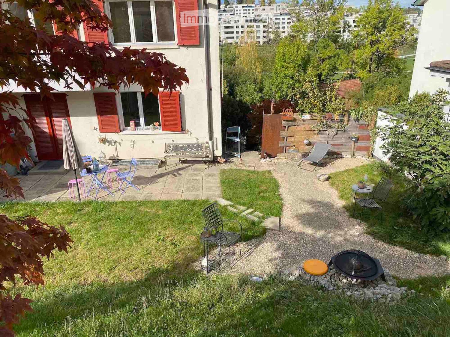Garden with a small patio area, seating, and a fire pit. The garden includes a grassy area, gravel pathways, and outdoor furniture.