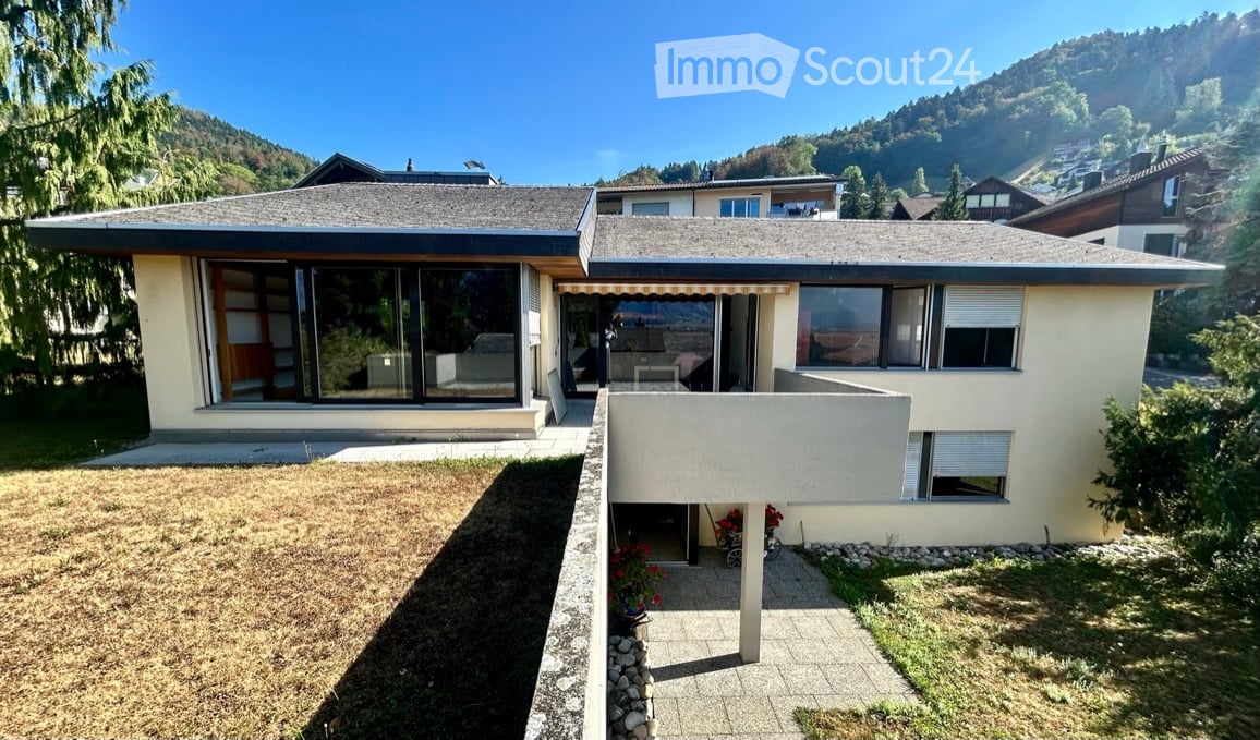 Two-story house with modern design, large windows, and a covered patio area. The house is surrounded by a well-maintained garden with a pathway leading to the entrance.