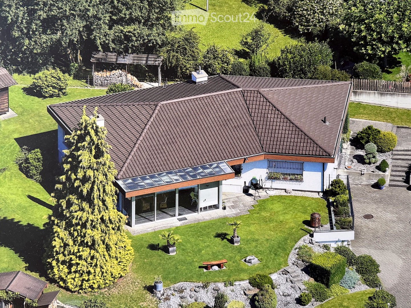 A two-story house with a tiled roof, surrounded by a well-maintained garden with trees, shrubs, and a patio area with outdoor furniture.