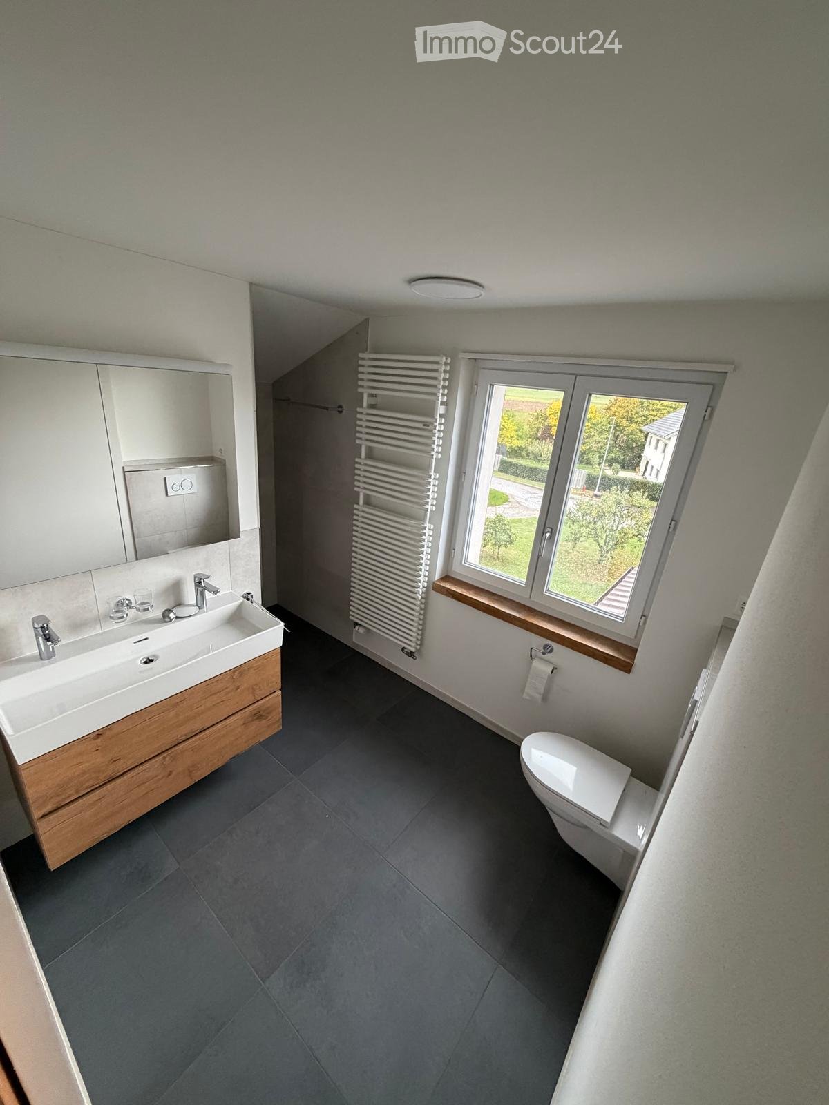 Modern bathroom with a double sink, wooden vanity, large mirror, radiator, and toilet. The room features dark grey tiles and a window with a view of the garden.