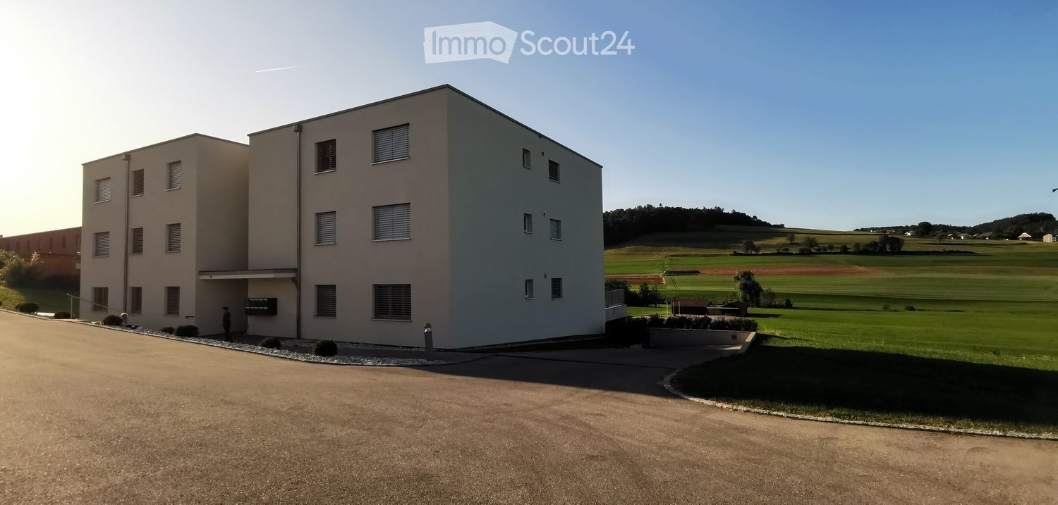Modern two-story building with light beige walls, multiple windows, and a flat roof, situated in a rural area with open fields and a clear blue sky.