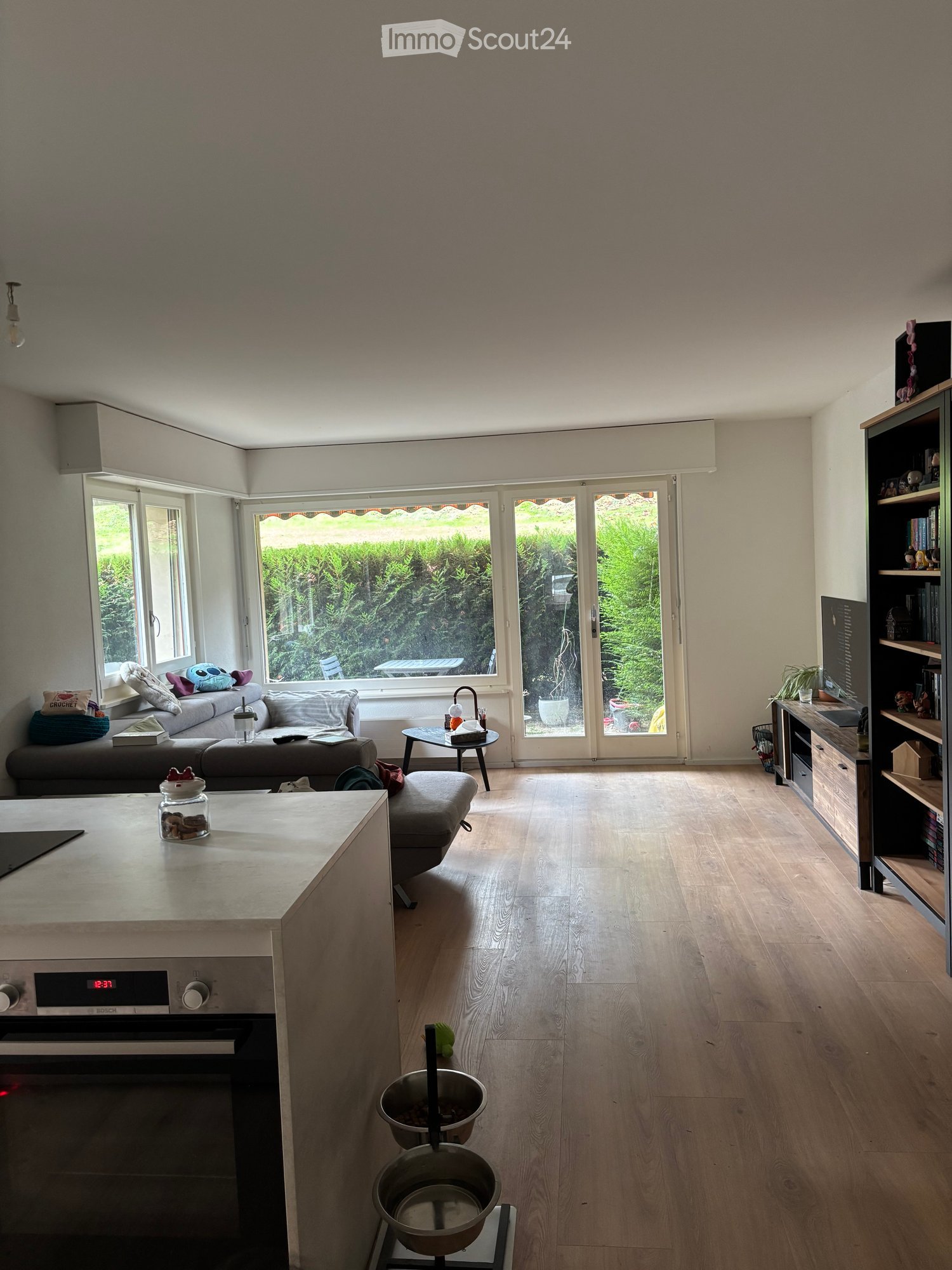 Open-plan living and dining area with light wooden flooring, large windows, and a modern kitchen island. Features a sofa, coffee table, small round table, and a bookshelf.