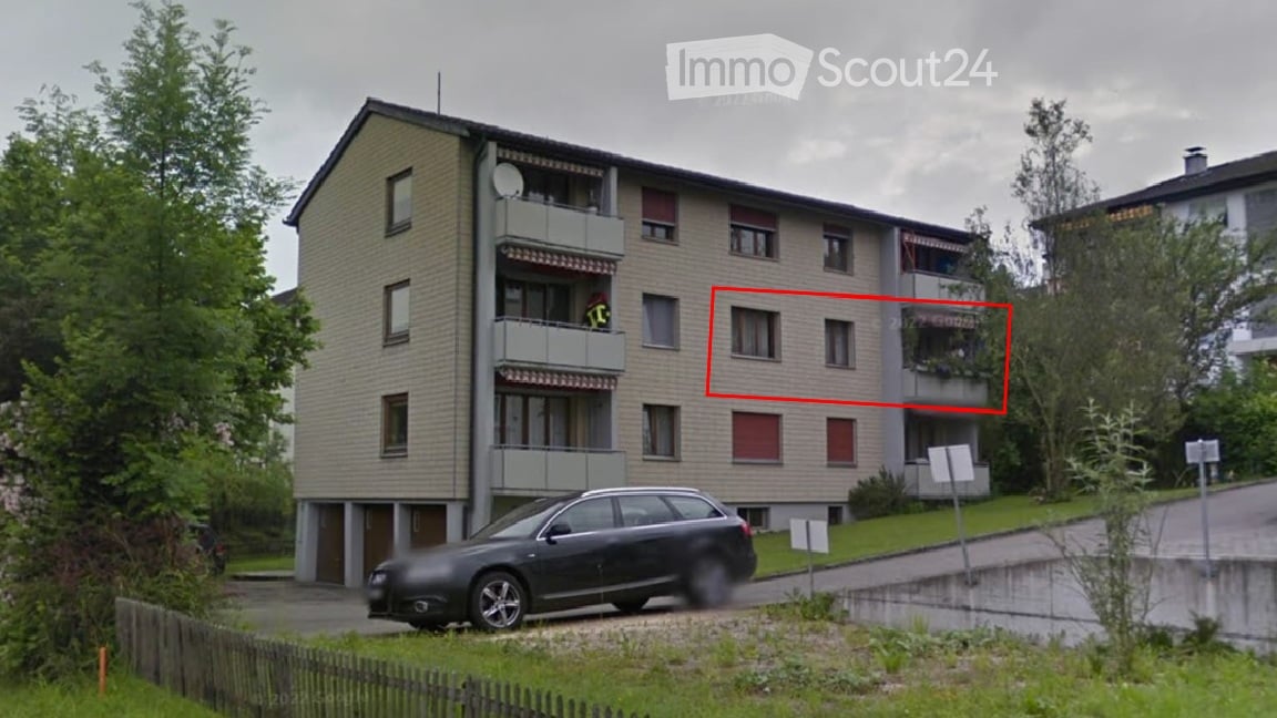 Four-story residential building with balconies, multiple windows, and a parked car in front.