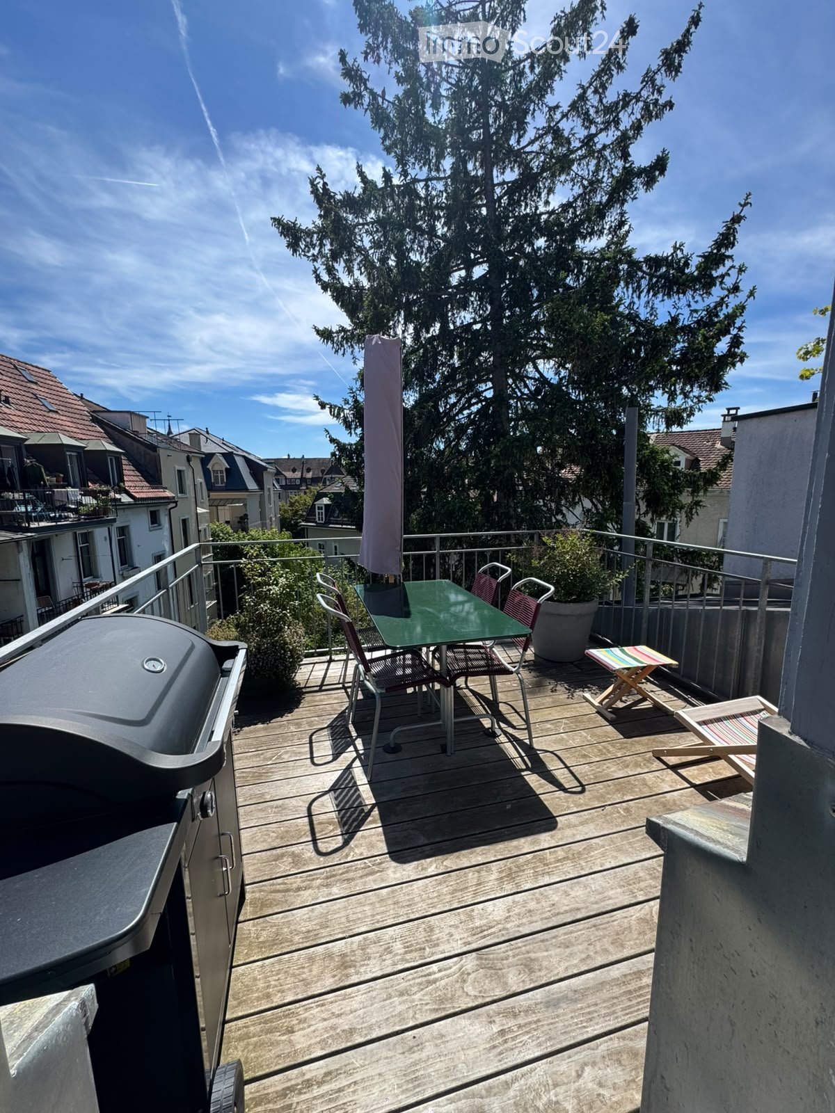 Wooden balcony with a table, chairs, umbrella, grill, and potted plants.