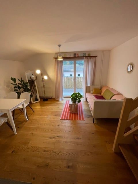 Bright living room with wooden flooring, large sliding glass door leading to a balcony, white sofa with colorful cushions, coffee table with a plant, dining area with white table and chairs, and a large mirror on the wall.