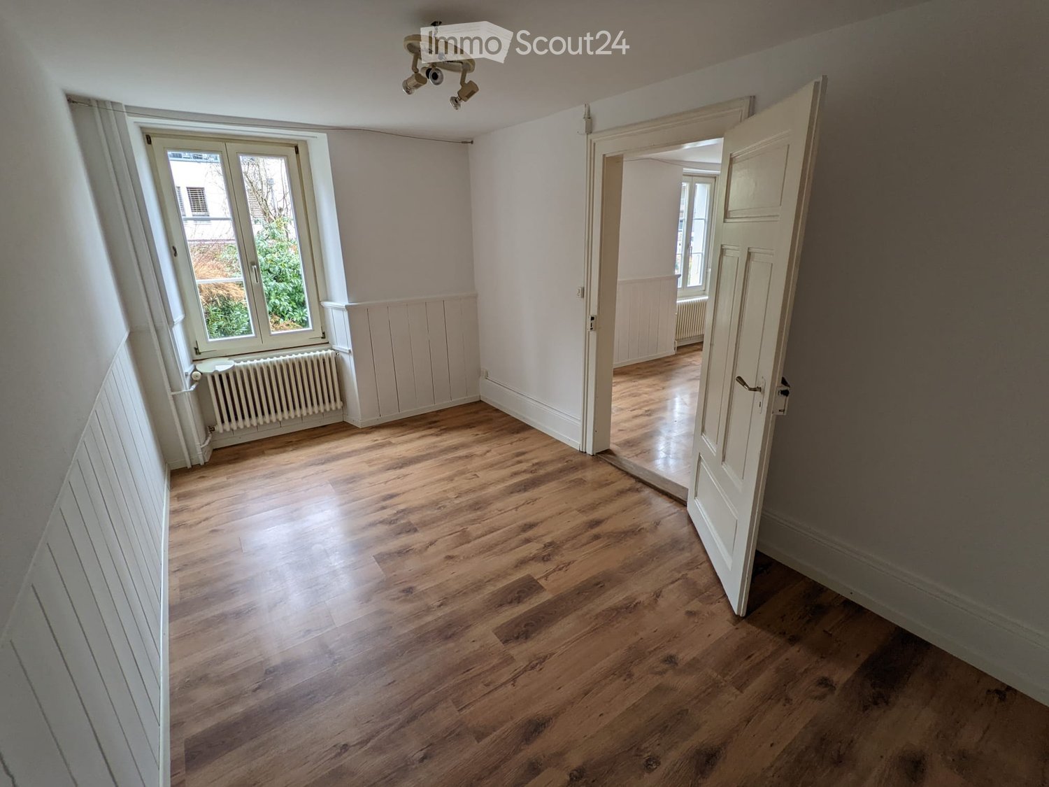 Empty room with wooden flooring, white walls, and a large window with a radiator below. An open door leads to another room with similar flooring and walls.