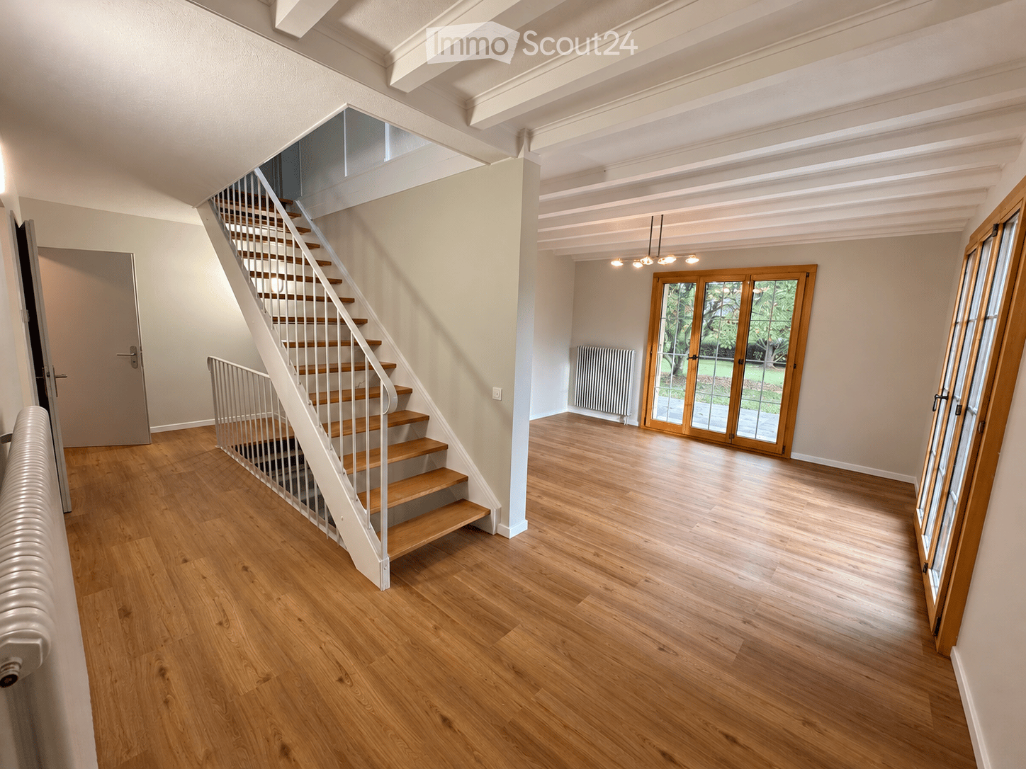 Spacious hallway with wooden flooring, a staircase leading upwards, and large windows providing natural light. The area is clean and well-lit, with a radiator visible on the left side.