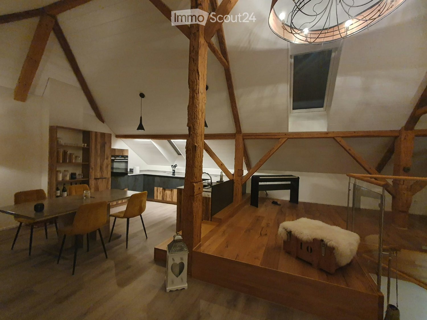 Open-plan attic space with wooden beams, a dining area, a kitchen area, and a small seating area with a fur-covered trunk. Exposed wooden beams and a skylight are visible.