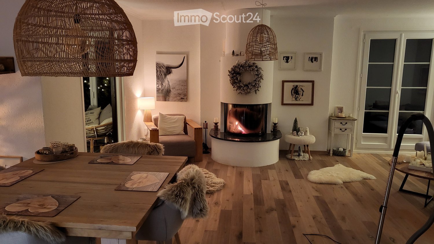 Cozy living and dining area with a central fireplace, wooden dining table, sofa, and decorative elements including a wreath and picture frames.