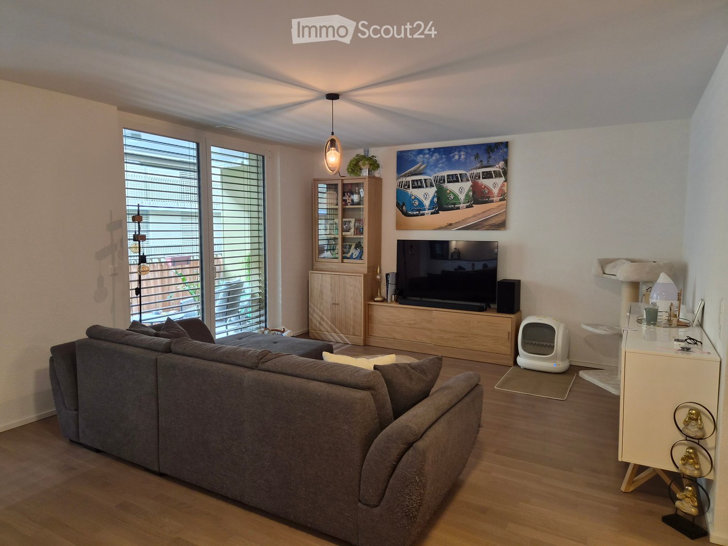 Modern living room with a grey sectional sofa, wooden coffee table, flat-screen TV on a wooden stand, cat tree, and a cat litter box. Large windows with blinds offer a view of the city.
