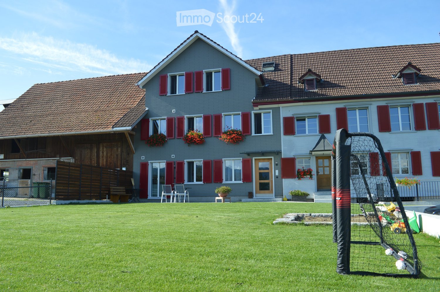 Two-story house with grey exterior, red shutters, and flower boxes. Features a well-maintained lawn, outdoor seating area, and a portable soccer goal.