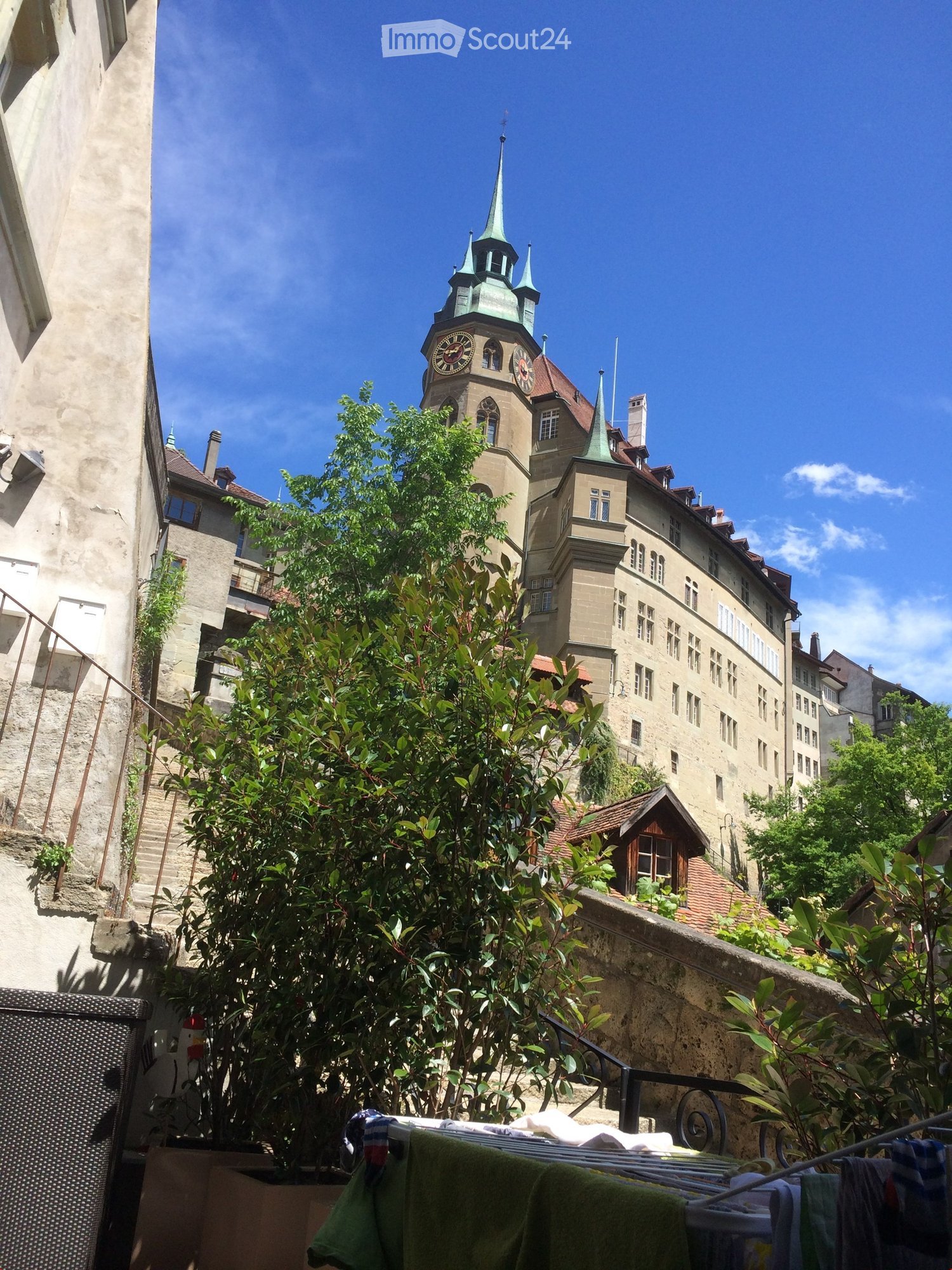 A historic building with a clock tower and green spire, surrounded by trees and greenery in a European city setting.