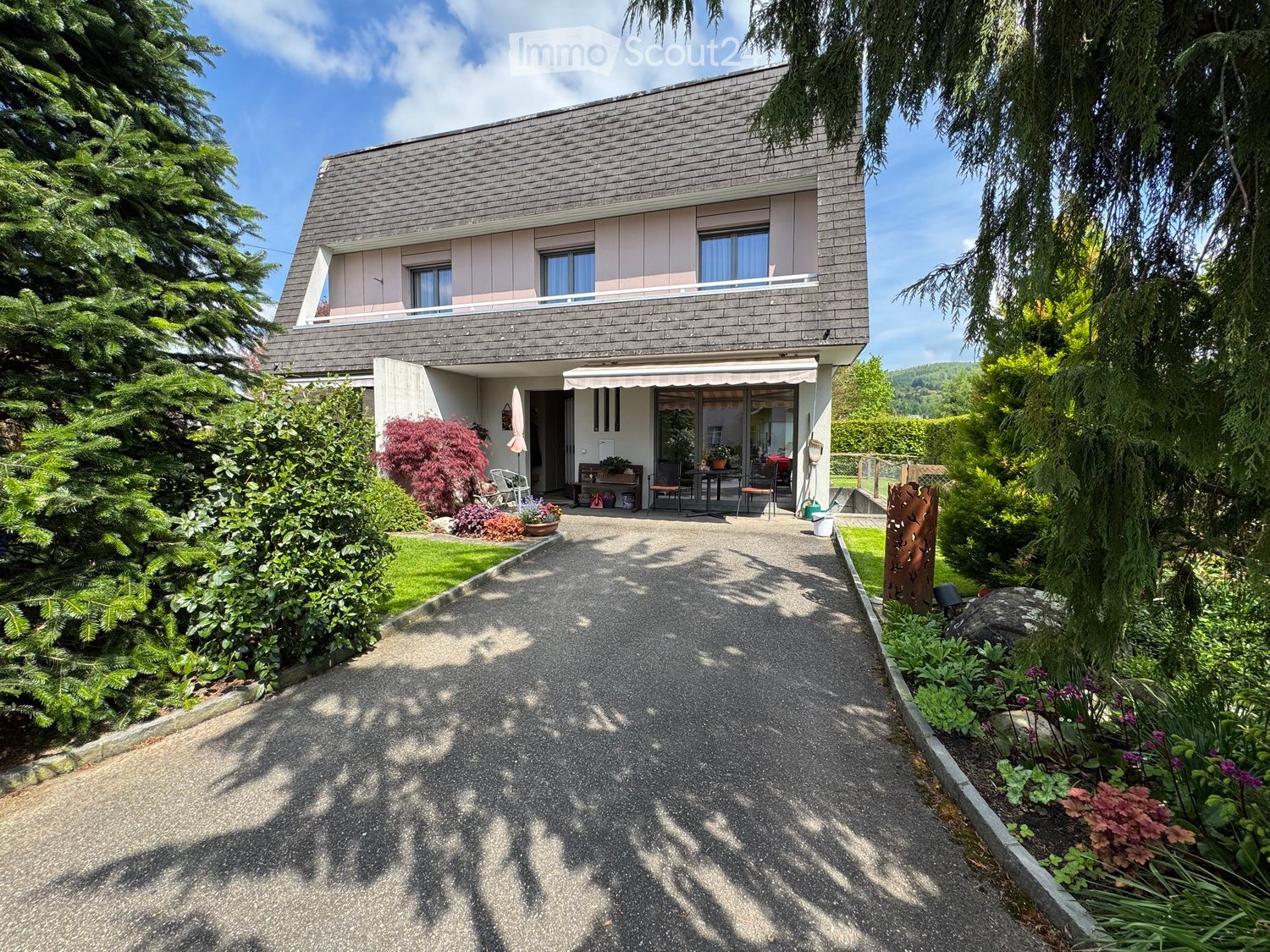 Modern two-story house with grey siding, large windows, and a covered entrance area. The house is surrounded by well-maintained gardens and trees.