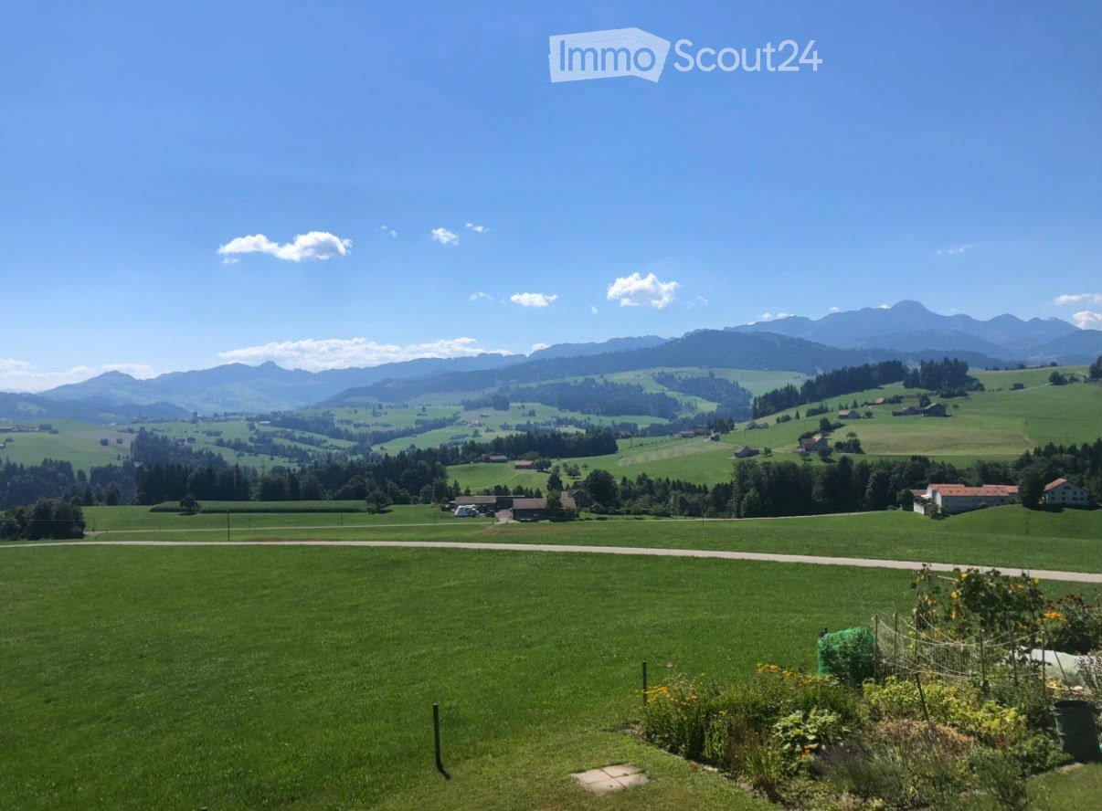 A panoramic view of a lush green valley with rolling hills, scattered houses, and distant mountains under a clear blue sky.