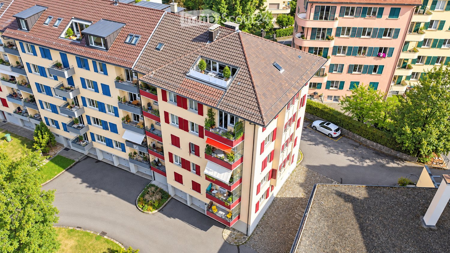 Aerial view of a multi-story residential building with balconies, surrounded by greenery and parking area.