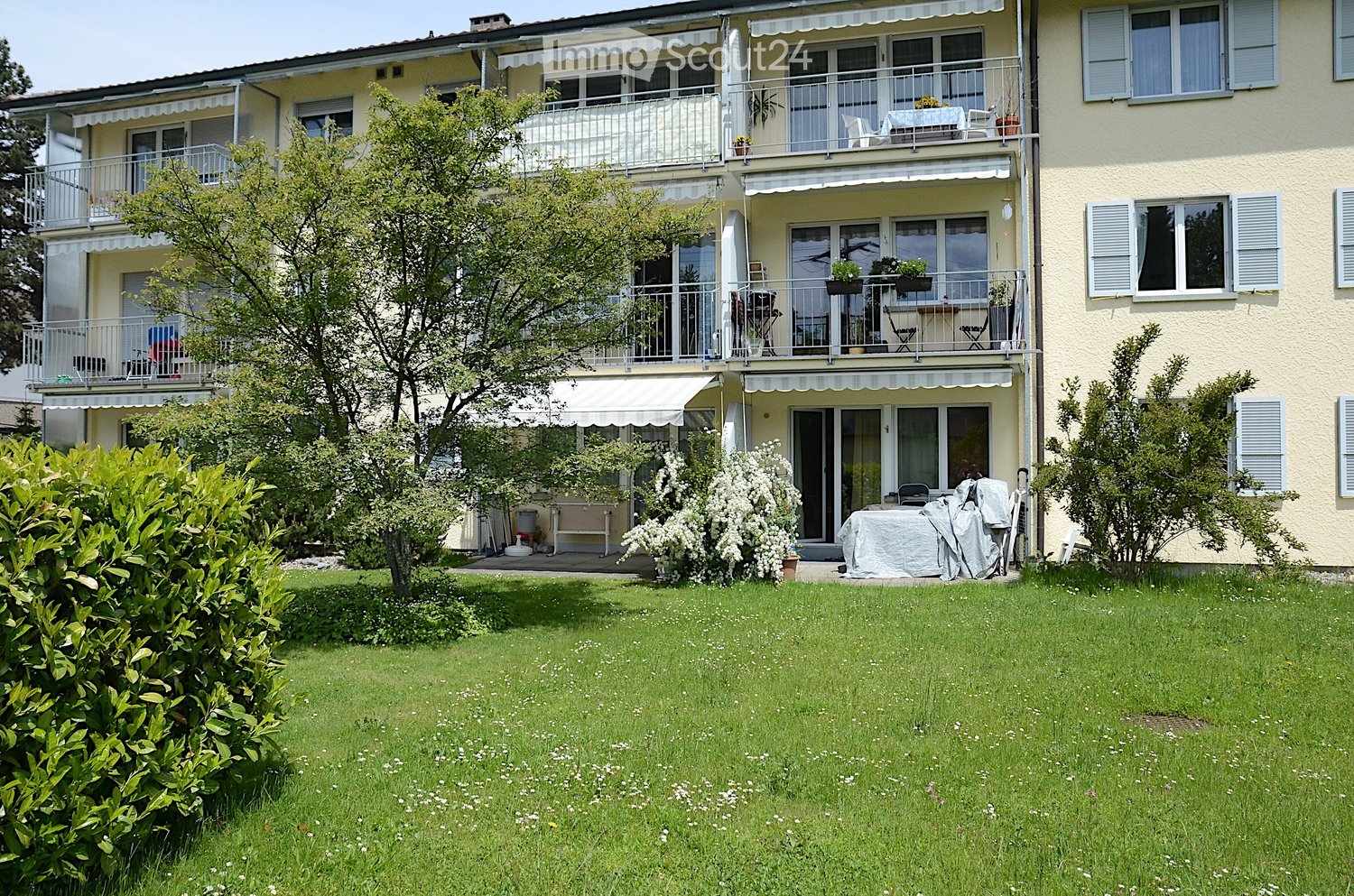 A multi-story residential building with balconies, awnings, and a well-maintained garden area with a covered patio and outdoor furniture.