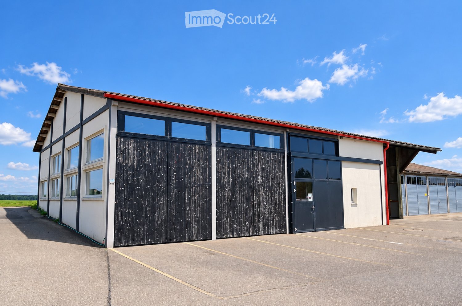 Modern single-story building with white walls, large windows, and dark wooden garage doors. The structure features a red roof trim and a spacious paved parking area.
