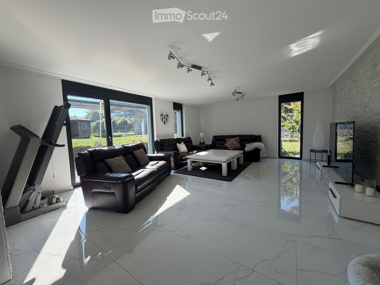 Modern living room with white marble flooring, large glass doors, leather sofas, coffee table, armchairs, ottoman, floor lamp, mirror, and fitness bike.