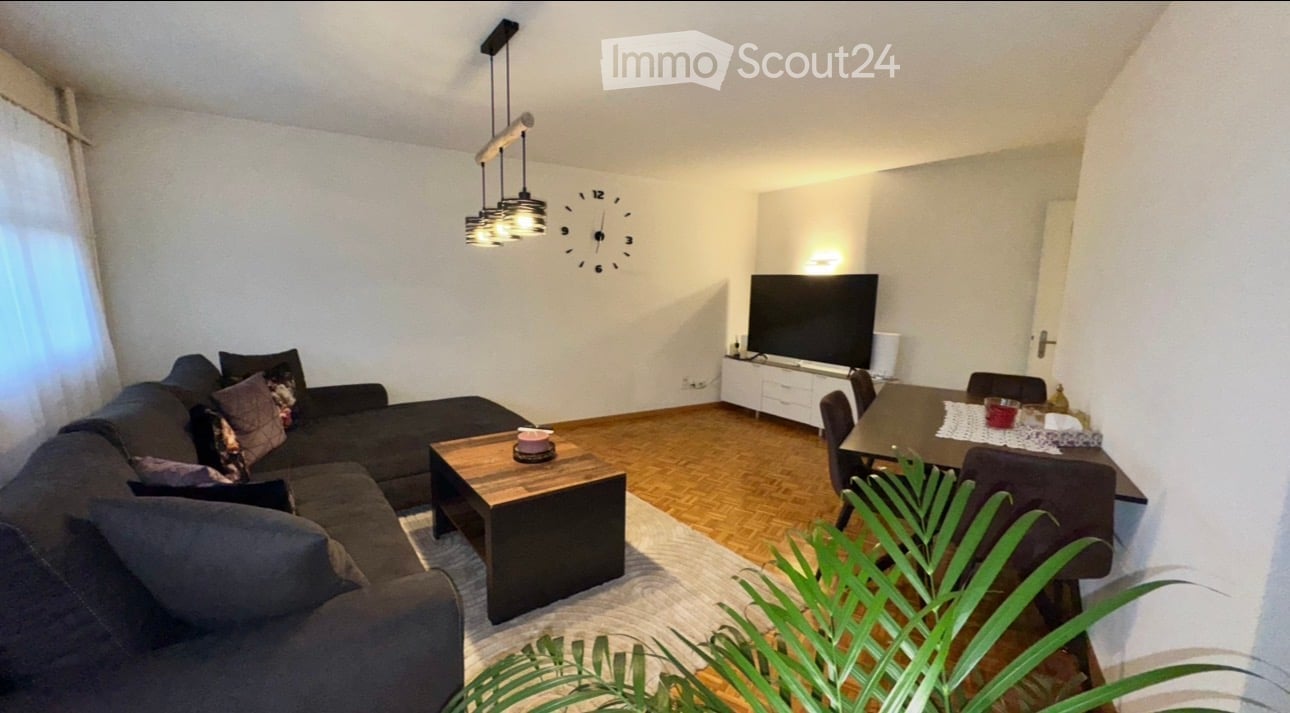 Modern living room with a dark grey sectional sofa, wooden coffee table, dining table with chairs, flat-screen TV, and a wall-mounted clock. The space is well-lit with pendant lights and features a large leafy plant in the foreground.