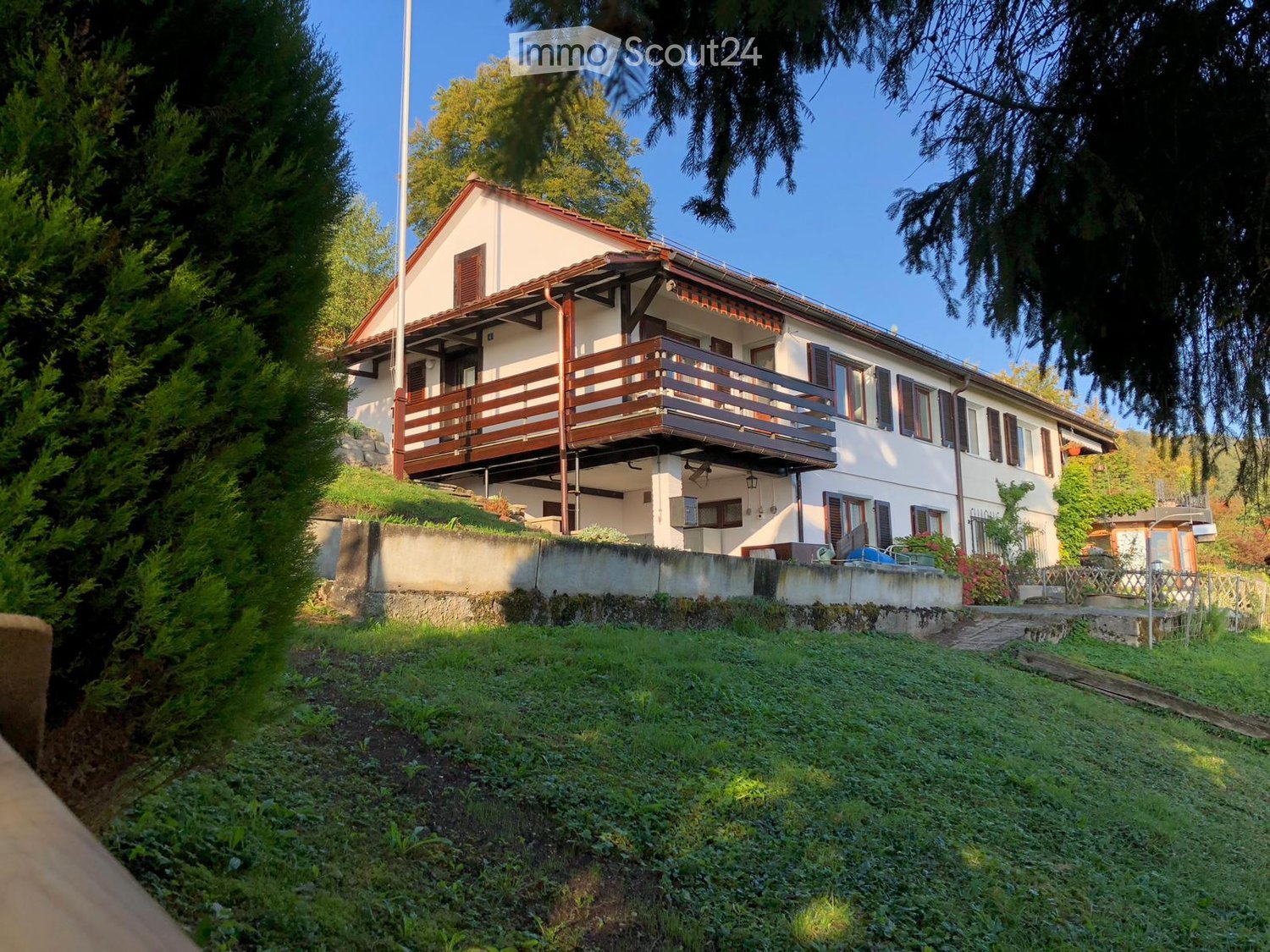Two-story house with a red-tiled roof, white exterior, and a wooden balcony on the upper level. The house is surrounded by greenery and has a raised ground floor.