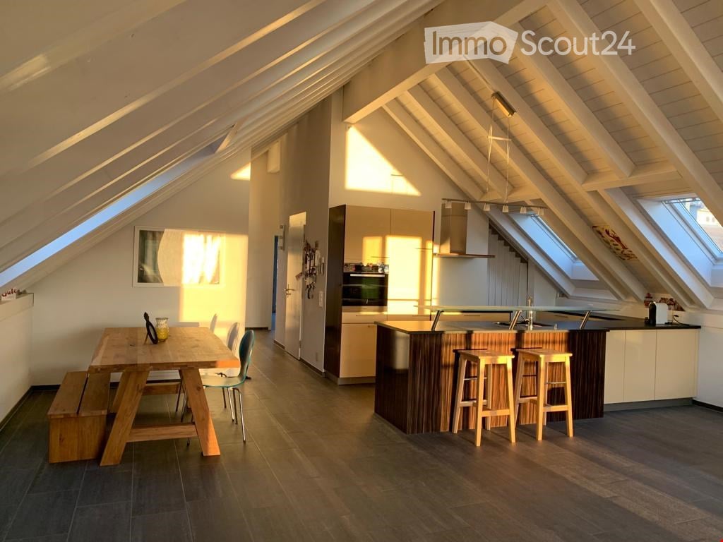 Bright attic space with wooden beams, featuring a kitchen area with an island, stools, and a dining table with chairs and a bench.