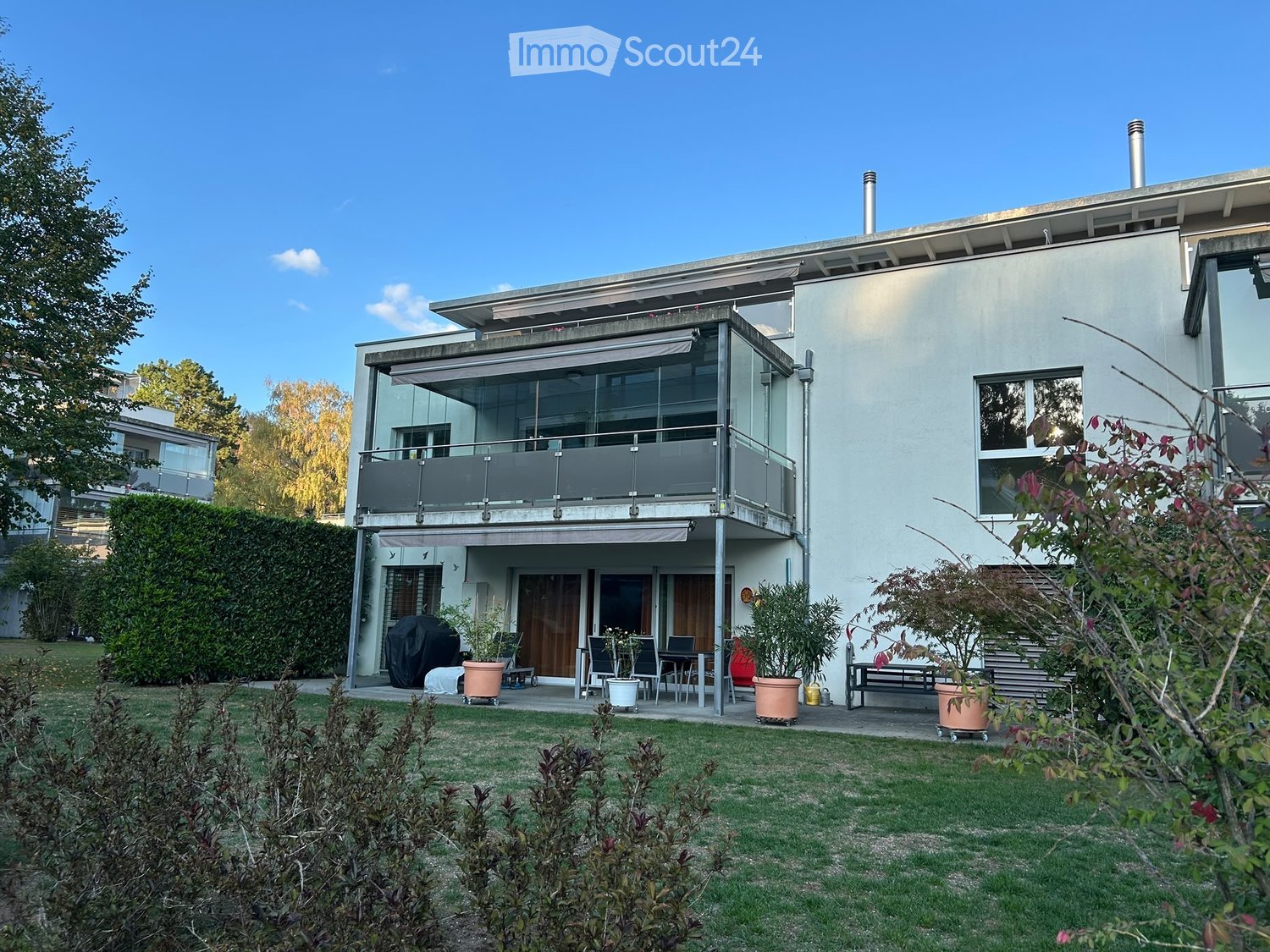 Modern two-story house with a balcony, large windows, and a well-maintained garden with potted plants and a bench.