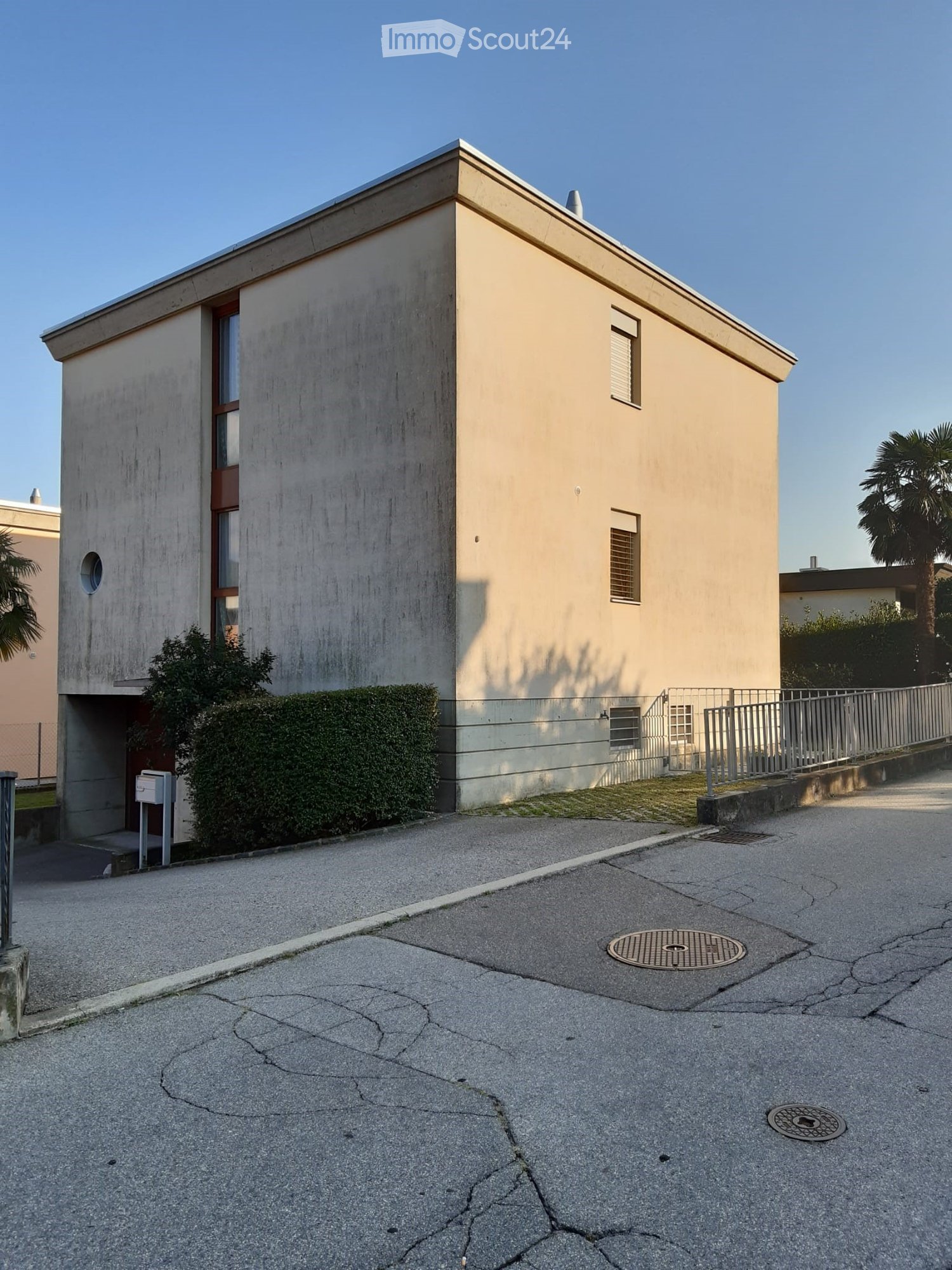 Modern two-story building with a flat roof, light beige facade, and small windows. The entrance features a small porch with a metal railing and a small garden area with trimmed hedges.