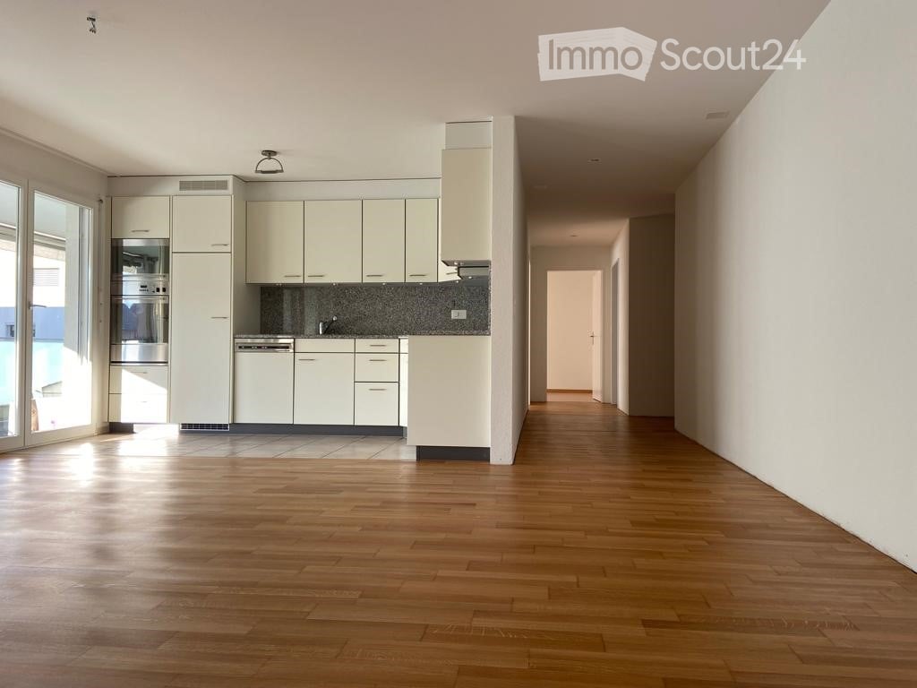 Empty room with wooden flooring, white walls, and a small kitchen area with cabinets and appliances visible in the background.