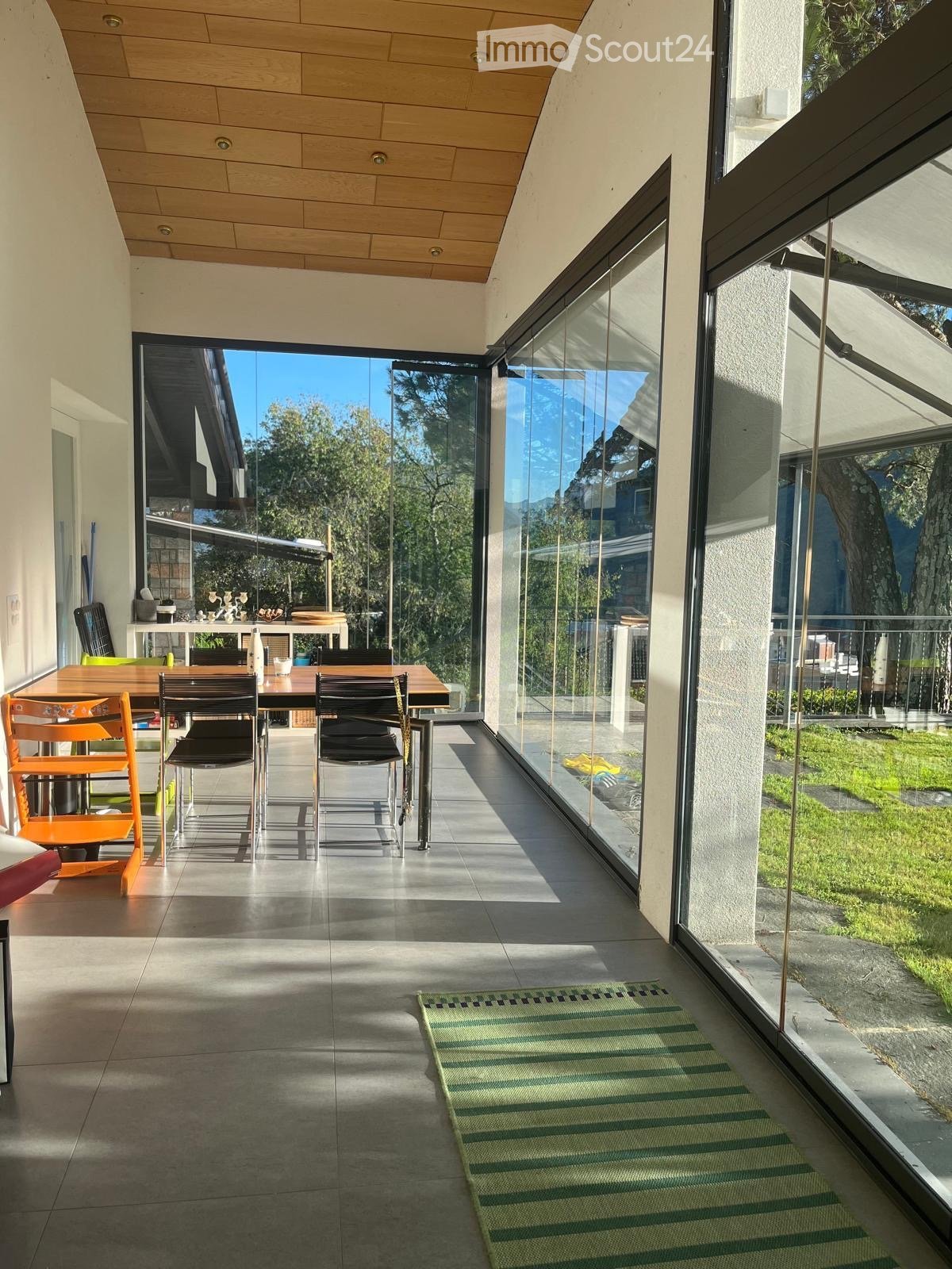 Bright, modern dining area with large glass walls, wooden table, and chairs, leading to a garden view.