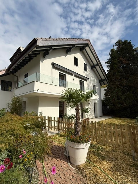 Modern two-story house with white exterior, large windows, and a balcony on the upper floor. The front yard features a wooden fence, potted plants, and a palm tree.