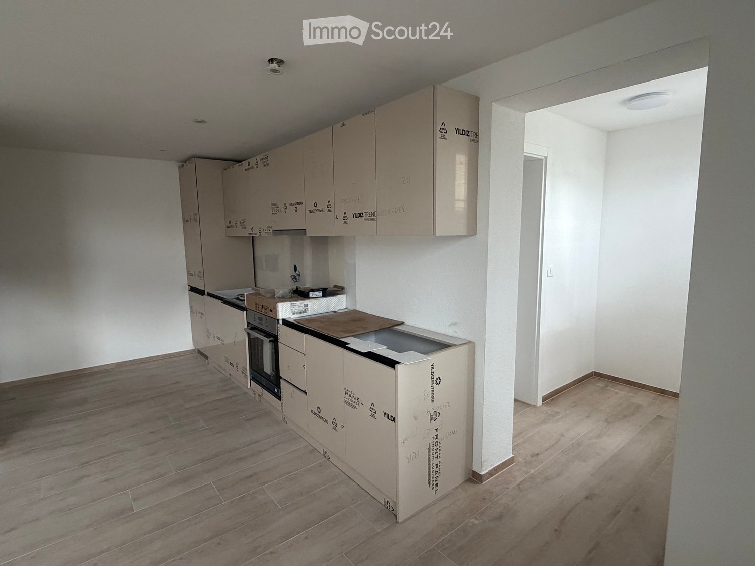 Empty kitchen with wooden flooring, white walls, and cardboard boxes stacked against the walls. No furniture or appliances are present.
