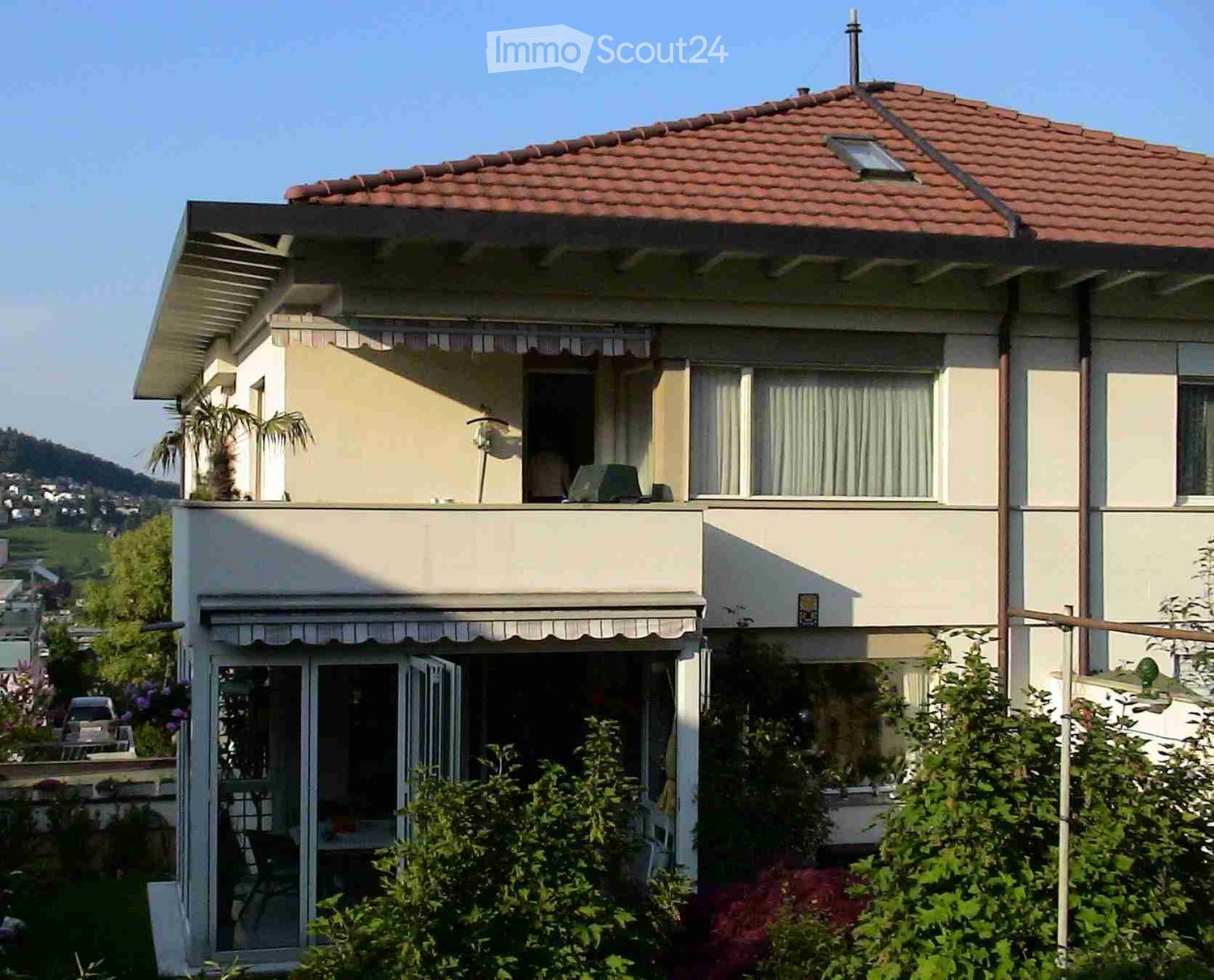 Two-story house with a red-tiled roof, beige exterior, large windows, and a balcony on the upper level. The house is surrounded by greenery and has a covered entrance area.
