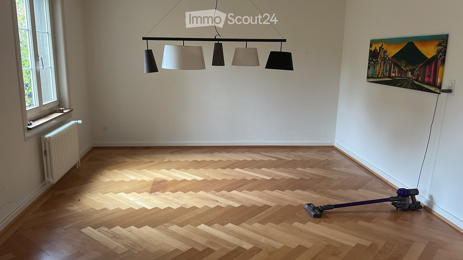 Empty room with wooden flooring, white walls, large window, radiator, and a painting on the wall. A vacuum cleaner is placed on the floor.