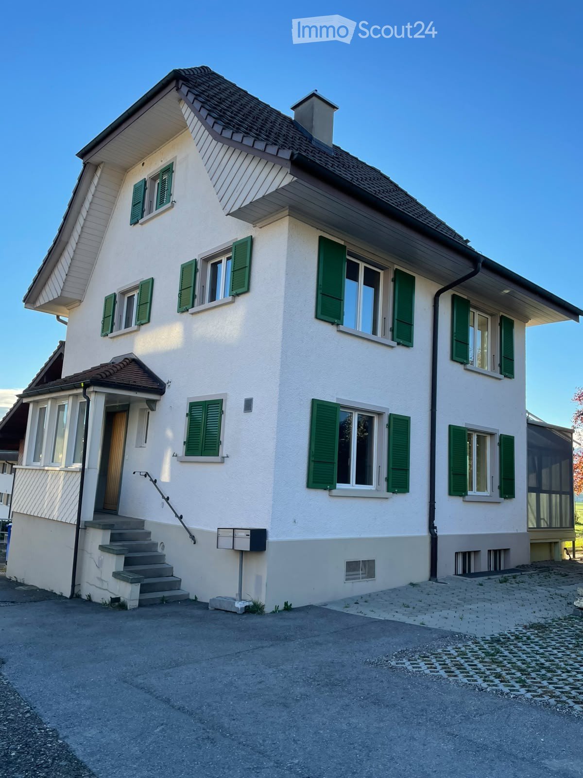 Two-story house with white walls, green shutters, and a brown tiled roof. Features include a front staircase, mailbox, and a small porch area.