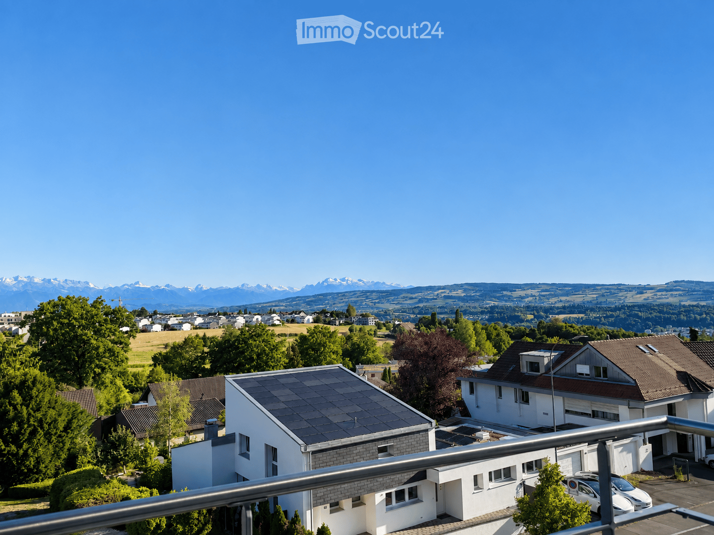 View from a balcony overlooking a suburban neighborhood with houses, trees, and distant mountains under a clear blue sky.