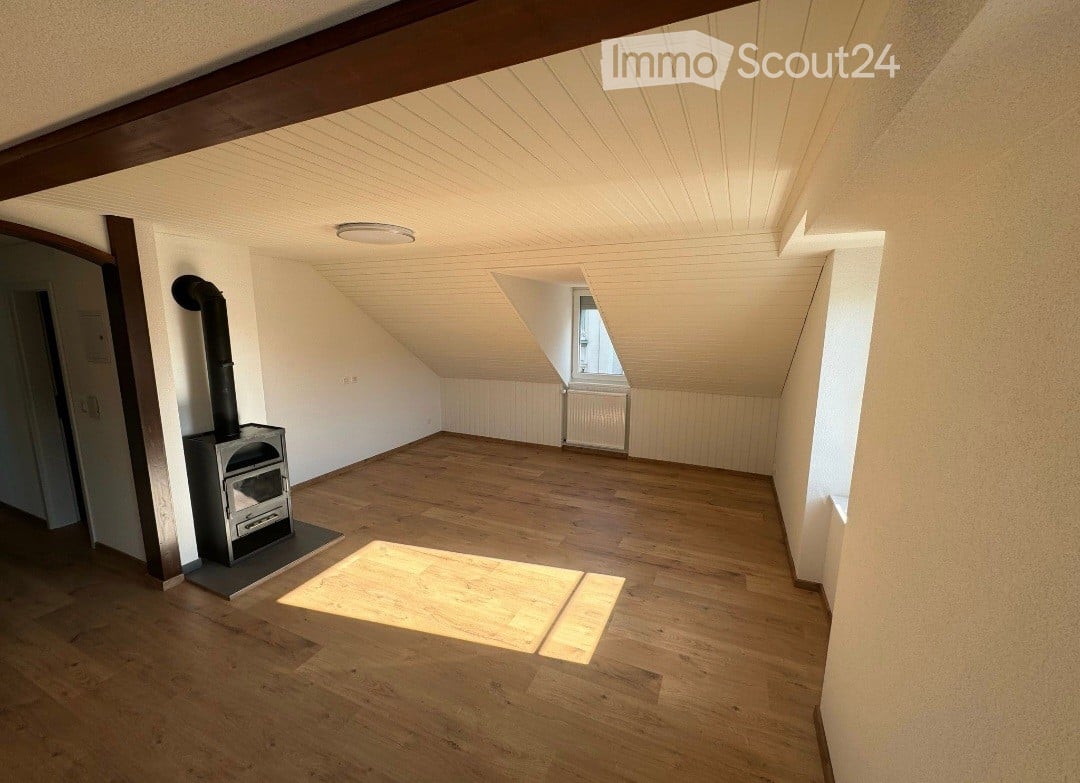Empty attic room with wooden flooring, exposed wooden beams, and a small wood-burning stove. Natural light streams in from a dormer window.