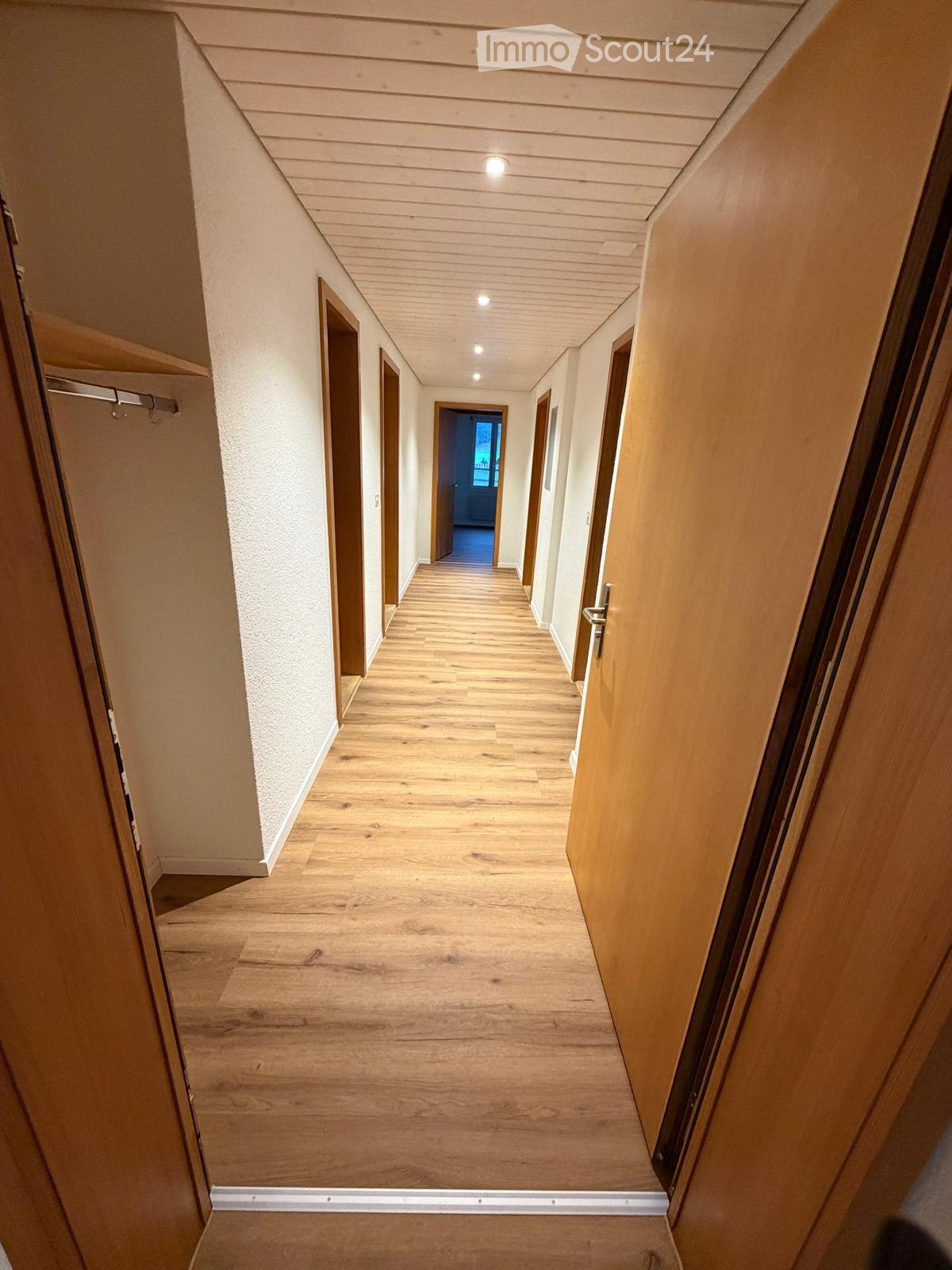 A well-lit hallway with wooden flooring, white walls, and several doors on both sides. The ceiling has recessed lighting, and the hallway appears clean and organized.