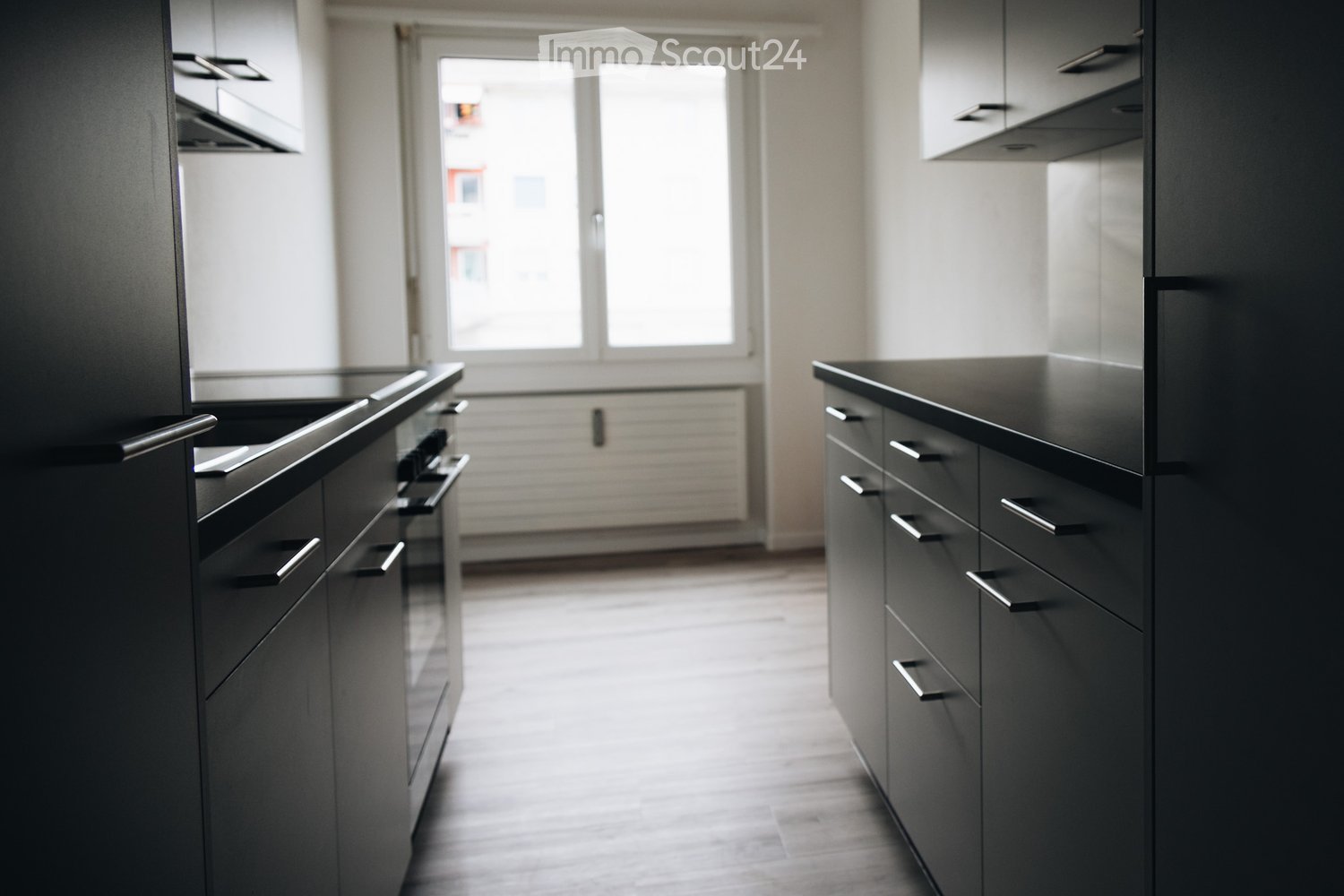 Modern kitchen with dark grey cabinets, stainless steel handles, and a sleek stove. The space features a large window allowing natural light to fill the room.