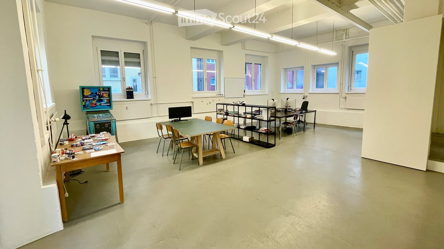 A spacious, well-lit room with multiple tables, chairs, shelves, a screen, and an arcade machine. The room has large windows and a clean, minimalist design.