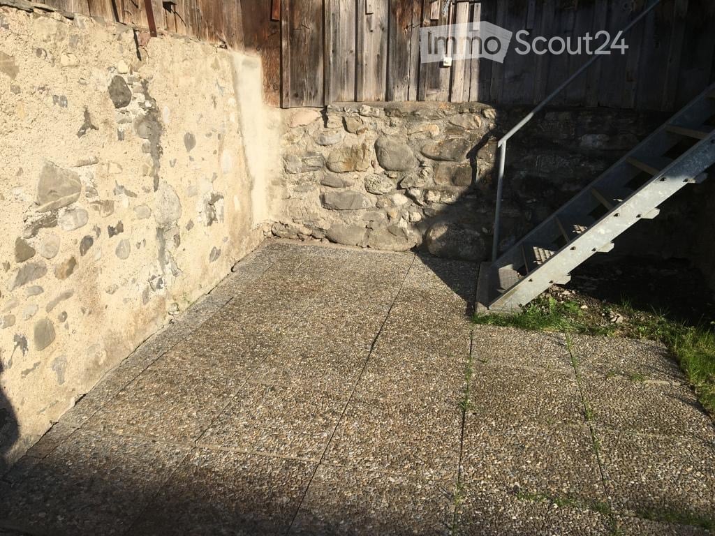 Raised concrete ground floor area adjacent to a stone wall, with a metal staircase leading up to a wooden structure.