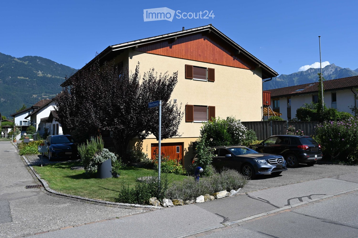 Two-story house with beige walls, red wooden roof, multiple windows with shutters, and a well-maintained garden with shrubs and flowers. Three cars are parked in the driveway.