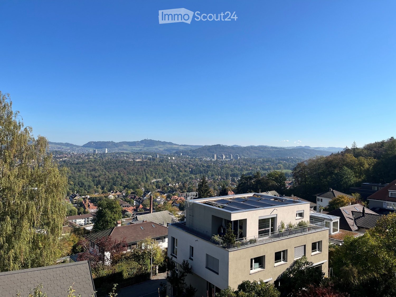 Modern multi-story house with a flat roof, large windows, and solar panels on the roof, surrounded by trees and a scenic cityscape in the background.