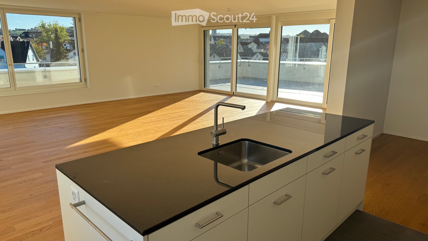 Empty kitchen with wooden flooring, black countertop, and stainless steel sink. Large windows and sliding doors lead to a balcony.