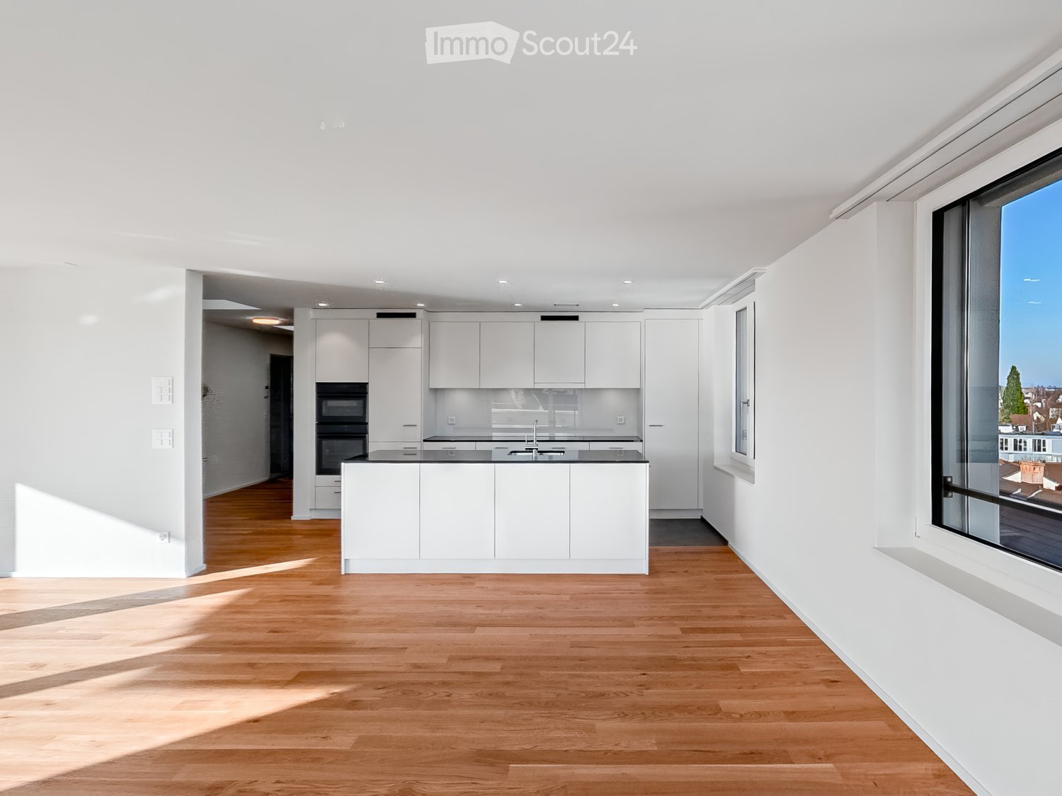 Modern kitchen with white cabinets, a central island, and a large window offering a view. The space features light wooden flooring and ample natural light.